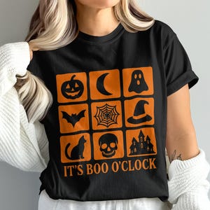 Op de afbeelding: Zwart T-shirt met oranje Halloween-thema graphics in een rasterpatroon. De graphics omvatten een pompoen, maan, spook, vleermuis, spinnenweb, heksenhoed, kat, schedel en spookhuis. De tekst "IT'S BOO O'CLOCK" is eronder gedrukt.