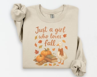 May include: Beige sweatshirt with the text "Just a girl who loves fall." The graphic includes pumpkins, fall leaves, a stack of books, and a duck wearing a scarf. The sweatshirt is a crew neck.