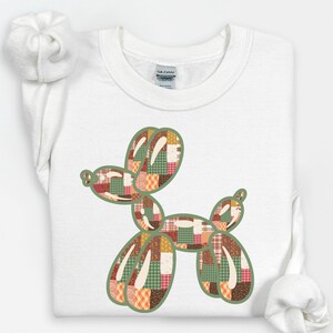 Könnte beinhalten: Weißes Sweatshirt mit einem Patchwork-Ballonhund-Design. Der Hund ist grün umrandet und mit verschiedenen Mustern in Rot-, Orange-, Grün- und Brauntönen gefüllt. Das Sweatshirt hat einen Rundhalsausschnitt und lange Ärmel.