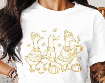May include: White t-shirt featuring a line drawing of three geese wearing sweaters, hats, and scarves. One goose holds a mug of coffee. Pumpkins and leaves are also depicted. A fall-themed graphic.