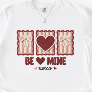 May include: White t-shirt with a Valentine's Day design. The design includes three rectangular frames with a heart and bows, the words "BE MINE", and "xoxo". The shirt is made of a soft material.