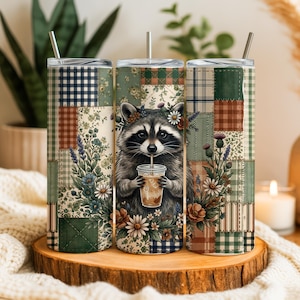 May include: Three stainless steel insulated tumblers with a patchwork design. Each tumbler features a different pattern, including plaid, floral, and solid colors. The center tumbler has a raccoon illustration drinking from a cup. The tumblers are on a wooden surface.