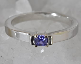Puede incluir: Un anillo de plata con una piedra preciosa rectangular de color azul oscuro. La piedra preciosa está engastada en un bisel dorado. La banda del anillo es de plata pulida. El anillo se muestra sobre una superficie blanca texturizada.