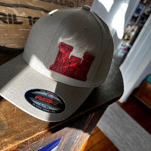 May include: A gray baseball cap with a red embroidered castle logo. The cap has a black Flexfit label with the text "The Original Flexfit L/XL It Fits Your Life".