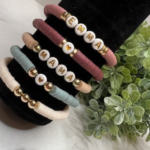 Peut inclure: Cinq bracelets faits à la main avec des perles plates colorées et des accents dorés. L'un des bracelets épelle "EMMA", un autre "MAMA". Les bracelets sont exposés sur un présentoir noir avec un feuillage vert en arrière-plan. Les bracelets sont dans les tons de rose, beige et bleu.