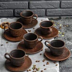 Peut inclure: Un ensemble de six tasses à café et soucoupes en céramique marron rustique. Les tasses ont une surface texturée et des anses incurvées. Les soucoupes sont rondes et complètent les tasses. Des grains de café sont dispersés.