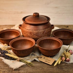 Può includere: Un set di pentole in ceramica marrone rustica, tra cui una pentola con coperchio e quattro ciotole, disposte su un panno di lino. La pentola e le ciotole hanno un aspetto strutturato e terroso. Sono presenti fiori secchi e un'etichetta "Welcome Home".
