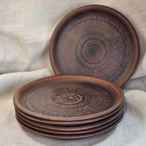 Puede incluir: Un conjunto de seis platos de cerámica marrón rústicos. Cinco platos están apilados, con un plato individual detrás. Los platos presentan un patrón circular y una superficie texturizada. Se muestran sobre una tela de tono neutro.