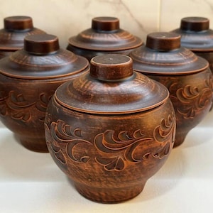 Op de afbeelding: Zes bruine keramische potten met deksels. Elke pot heeft een afgeronde body en een deksel met een kleine knop. De potten zijn versierd met een bloemmotief en zijn in een groep gerangschikt.