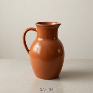 May include: A terracotta-colored ceramic pitcher with a curved handle. The 2.5-liter pitcher has a rounded body and a slightly flared rim. The glossy finish reflects light, highlighting its smooth surface. The pitcher sits on a light-colored surface.