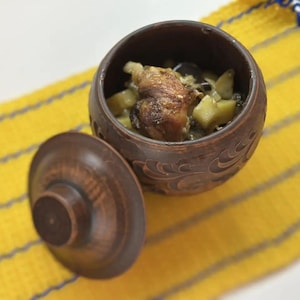 May include: A small, round, brown ceramic bowl with a lid, filled with cooked food. The bowl sits on a yellow and blue striped cloth. The lid is off to the side, revealing the contents of the bowl.