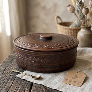 May include: A brown ceramic casserole dish with a lid, featuring a floral design around the rim and on the lid. The dish sits on a wooden table with a small spoon and a tag that reads "THE HERB & SALT BOX".