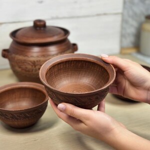 K&ouml;nnte beinhalten: Ein Set aus braunen Keramikschalen und einem Topf mit Deckel. Die Schalen haben ein strukturiertes, geschnitztes Design am Rand. Der Topf hat einen Deckel und Griffe. Die T&ouml;pferwaren werden auf einer hellen Holzoberfl&auml;che pr&auml;sentiert.