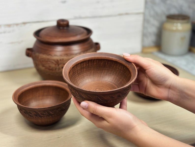 K&ouml;nnte beinhalten: Ein Set aus braunen Keramikschalen und einem Topf mit Deckel. Die Schalen haben ein strukturiertes, geschnitztes Design am Rand. Der Topf hat einen Deckel und Griffe. Die T&ouml;pferwaren werden auf einer hellen Holzoberfl&auml;che pr&auml;sentiert.