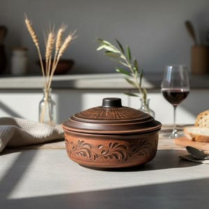 以下が含まれることがあります： 蓋付きの茶色のセラミックキャセロール皿。浮き彫りの花柄が特徴です。皿は、赤ワインのグラス、パン、リネンのナプキンが置かれたテーブルの上にあります。キッチンには、装飾用の小麦の穂が含まれています。