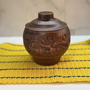 May include: A brown ceramic jar with a lid, featuring carved floral designs. The jar sits on a yellow and blue striped woven mat. The jar has a round shape and a small knob on the lid.