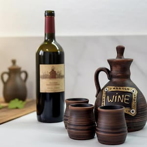 Op de afbeelding: Een wijnfles met etiket, een bruine keramische kan met een handvat en het woord "WINE", en drie kleine bruine keramische bekers. De items zijn gerangschikt op een wit oppervlak, wat wijst op een wijnproeverij.