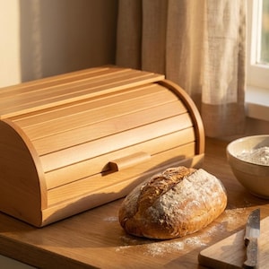 May include: A light-coloured wooden bread bin with a roll-top lid is on a wooden surface, next to a loaf of bread and a chopping board. A bowl of flour and a knife are also on the counter. The scene is lit by sunlight.