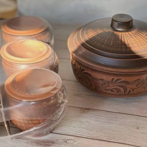 May include: A collection of brown ceramic bowls with lids, some individually wrapped in clear plastic. The bowls have a rustic, handcrafted appearance with a decorative floral pattern on the sides. The lids have a small knob.