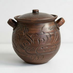 Könnte beinhalten: Ein brauner Keramiktopf mit Deckel und zwei Griffen. Der Topf hat geschnitzte Blumenmuster am Körper. Der Deckel hat einen kleinen Knopf. Der Topf dient wahrscheinlich zum Kochen oder Aufbewahren. Maße in Zentimetern.