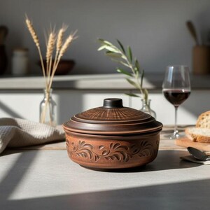 May include: A brown ceramic casserole dish with a lid, featuring an embossed floral design. The dish is on a table with a glass of red wine, bread, and a linen napkin. The dish measures approximately 10 inches in diameter.