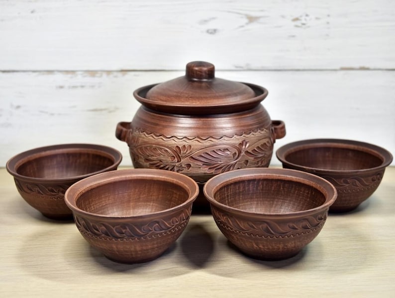 K&ouml;nnte beinhalten: Ein Set aus braunen Keramik-Servierschalen und einem Topf mit Deckel. Die Schalen und der Topf haben ein rustikales Aussehen mit geschnitzten Blumen- und Blattmustern. Das Set enth&auml;lt einen gro&szlig;en Topf mit Griffen und vier kleinere Schalen.