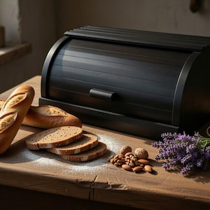May include: A black wooden bread box with a roll-top lid sits on a wooden table. Sliced bread, baguettes, nuts, and a bundle of lavender are arranged nearby. The bread box has a handle and a curved design.