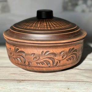 Peut inclure: Un pot en céramique marron avec un couvercle. Le pot a une forme ronde avec un motif floral décoratif sculpté autour de la base. Le couvercle est marron foncé avec un bouton noir. Le pot est posé sur une surface en bois.