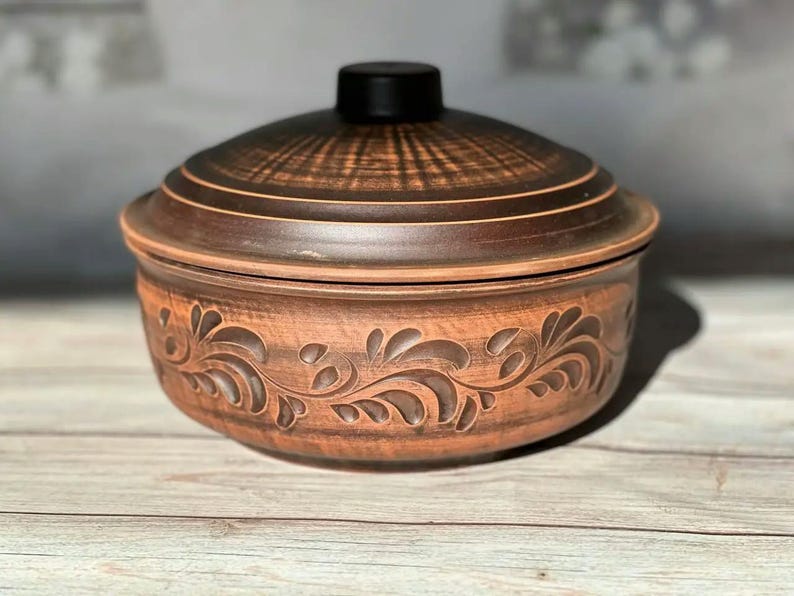 May include: A brown ceramic pot with a lid. The pot has a round shape with a decorative floral pattern carved around the base. The lid is dark brown with a black knob. The pot is sitting on a wooden surface.