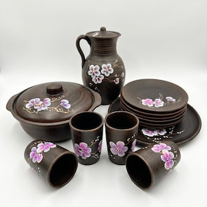 May include: A dark brown ceramic dinnerware set featuring a hand-painted pink and white cherry blossom motif. The collection includes a lidded pitcher, a lidded serving bowl, four cups, and a stack of six plates, all with a consistent floral design.