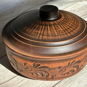 May include: A brown ceramic casserole dish with a lid. The lid has a dark brown knob and a textured pattern. The dish has a decorative floral design around the base. The dish is sitting on a wooden surface.