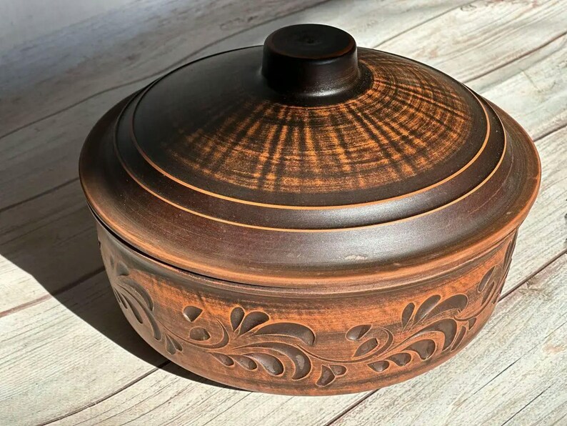 May include: A brown ceramic casserole dish with a lid. The lid has a dark brown knob and a textured pattern. The dish has a decorative floral design around the base. The dish is sitting on a wooden surface.