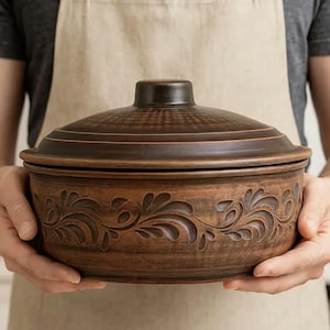 May include: A brown ceramic casserole dish with a lid, held by a person wearing an apron. The dish features a decorative floral pattern carved around the base. The lid has concentric rings and a small knob.