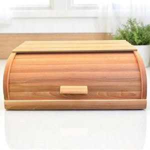 Puede incluir: Una panera de madera con tapa enrollable y asa rectangular. La panera es de madera de color claro y tiene un frente curvo. La panera está sobre una superficie blanca.