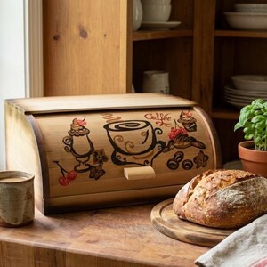 Peut inclure: Un coffre à pain en bois avec un couvercle roulant, décoré d'illustrations sur le thème du café. Une miche de pain est posée sur une planche à découper en bois, à côté d'une tasse de café. Une petite plante en pot et une fenêtre sont en arrière-plan.