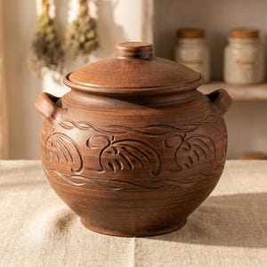 May include: A brown ceramic pot with a lid and handles. The pot has a rounded shape and is decorated with carved leaf and vine patterns. It is sitting on a beige linen surface.