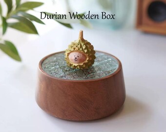 May include: A wooden music box with a small, carved durian fruit figurine on top. The durian has a cute, cartoon face and is painted in shades of green and yellow. The music box is made of dark wood and has a circular, patterned lid.