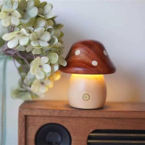 Pode incluir: Uma luz noturna de madeira em forma de cogumelo, com a parte superior marrom e bolinhas brancas. A base é de madeira clara e tem um pequeno botão amarelo na lateral.
