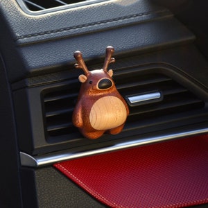Puede incluir: Un ambientador de coche de reno de madera marrón con vientre marrón claro y cuernos. El reno está colgado de una rejilla de ventilación del coche.