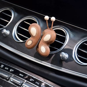 Op de afbeelding: Een houten autoluchtverfrisser in de vorm van een vlinder met witte accenten. De vlinder is bevestigd aan een ventilatieopening van een auto.