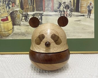 Puede incluir: Una caja de madera para guardar pequeños objetos con forma de panda, con tonos de madera marrón y clara. La caja es redonda y tiene una tapa que se abre para revelar un pequeño compartimento en el interior.
