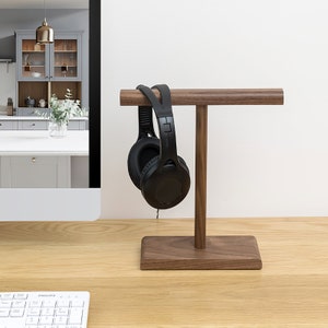 Puede incluir: Un soporte para auriculares de madera con un par de auriculares negros colgados. El soporte está hecho de madera de nogal y tiene un diseño simple y moderno.