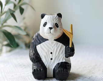 Puede incluir: Una figurita de panda de madera en blanco y negro con un palo de bambú dorado en la mano. El panda lleva un esmoquin y tiene una cara tallada.