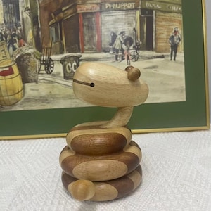 Puede incluir: Una serpiente de juguete de madera con un cuerpo segmentado. La serpiente está hecha de madera clara y oscura y tiene una cabeza marrón con dos pequeños ojos negros. La serpiente está enroscada en forma de espiral.