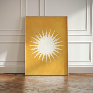 Puede incluir: Una impresión artística enmarcada con un diseño de sol blanco sobre un fondo amarillo. La obra de arte se exhibe en una habitación con molduras blancas y un suelo de madera. La ilustración del sol tiene una apariencia texturizada.