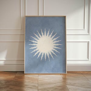 May include: A framed art print featuring a stylized sun design. The sun is depicted in a cream color against a light blue background. The artwork is encased in a natural wood frame, suitable for wall decor.