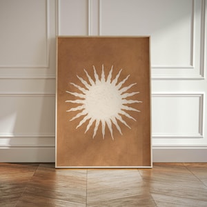 May include: A framed art print featuring a sun design. The sun is depicted in off-white with radiating lines against a brown background. The frame is a light wood color. The artwork is displayed against a white wall and wooden floor.