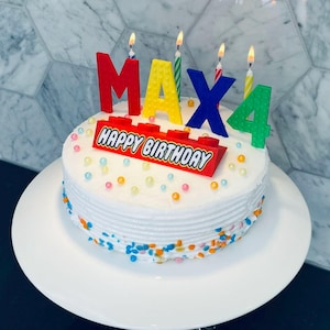 May include: A white cake with colorful sprinkles and a red, yellow, blue, and green Lego-themed Happy Birthday banner with candles that spell out "MAX 4".