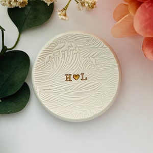 Puede incluir: Plato de cerámica blanco para anillos con diseño de olas y las iniciales "H♥L" en dorado.