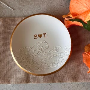 Pode incluir: Prato redondo de cerâmica branca com borda dourada e design de ondas. O prato apresenta as iniciais "B" e "T" com um coração dourado. Está sobre uma superfície de cor neutra.